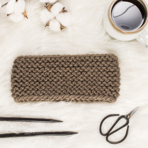 knit headband on a fur blanket with knitting needles, coffee & scissors