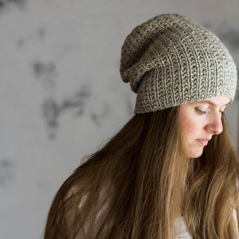 Hand knit super bulky slouchy hat in a soft neutral yarn, shown from the side and back on a woman with long hair, highlighting the textured rib stitches and rounded crown shaping.