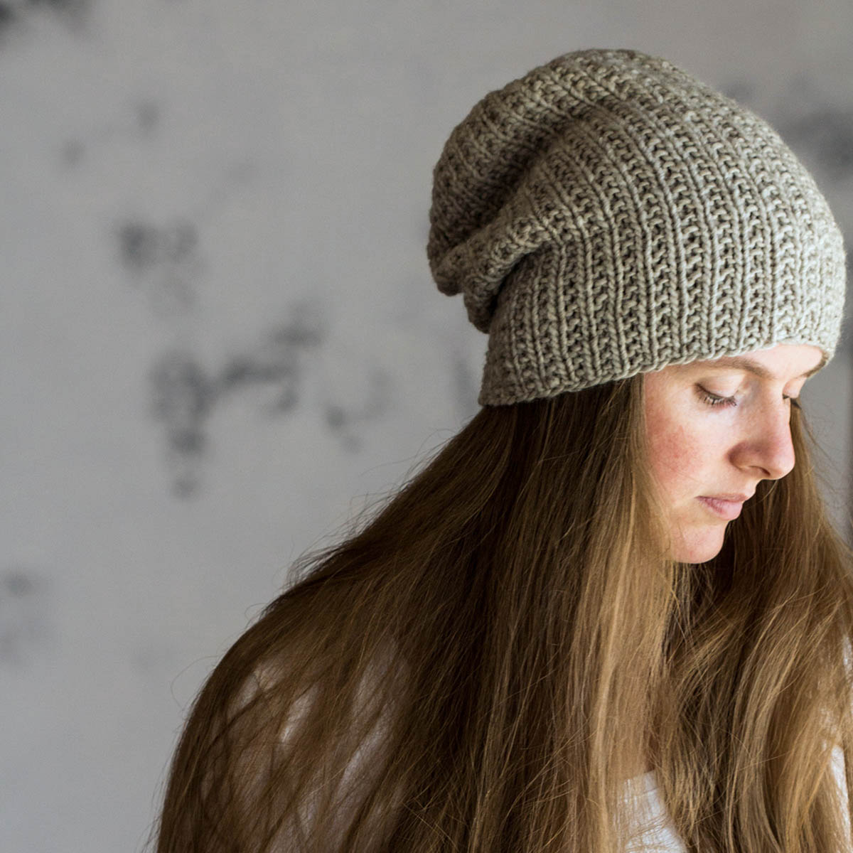 Hand knit super bulky slouchy hat in a soft neutral yarn, shown from the side and back on a woman with long hair, highlighting the textured rib stitches and rounded crown shaping.
