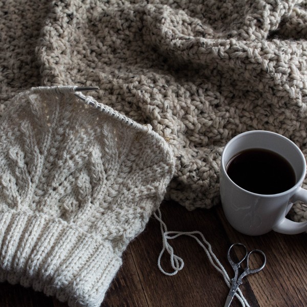 cozy scene of a hat partially knit on needles with coffee and a knit blanket