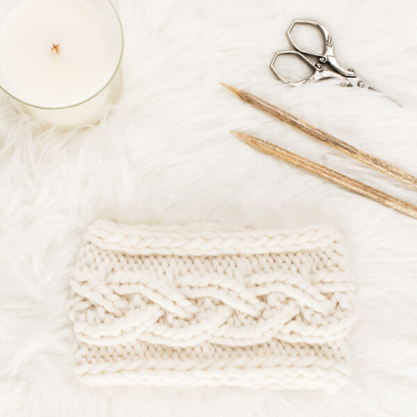 chunky knit cable knitting pattern on a fur blanket with a candle, knitting needles & scissors.