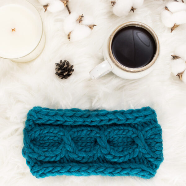 cozy scene of a cross cable knit headband on a fur blanket with coffee & a candle.