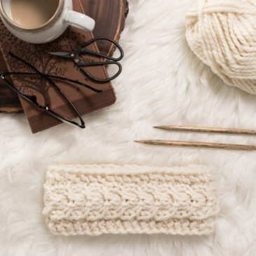 pic of a fish tail braid knitted headband on a faux fur blanket with coffee & knitting needles.