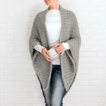 Woman wearing a chunky gray triangle shawl, holding a mug labeled "Grateful," standing against a white brick wall.