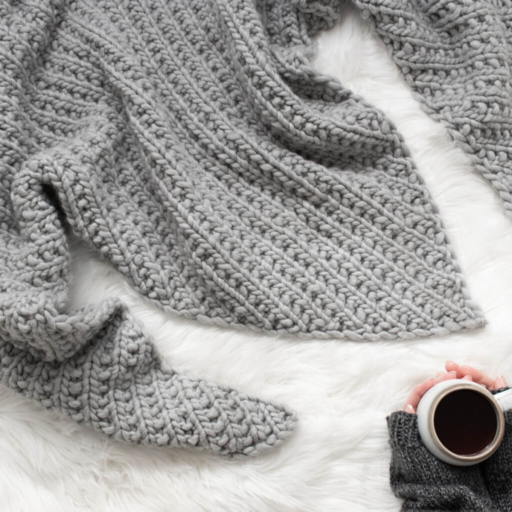 Chunky gray triangle shawl displayed on a white faux fur surface with a cup of coffee in hand.