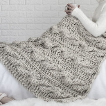 Model snuggled up in a cable knit blanket with a cup of coffee.