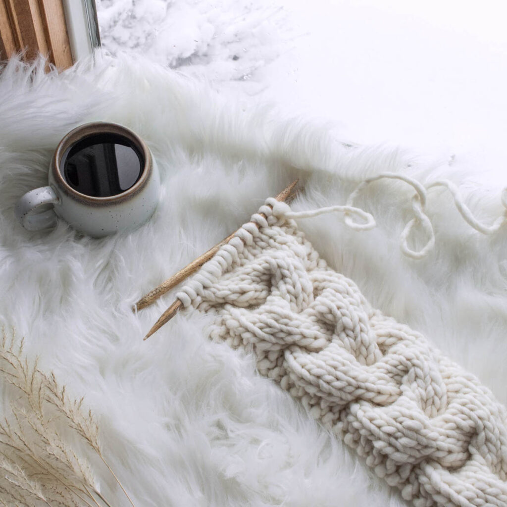 cable knit headband on a fur blanket with knitting needles & a coffee in a wintery window