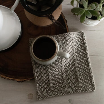knitted dishcloth on a wooden platter with a cup of coffee, coffee maker and plant