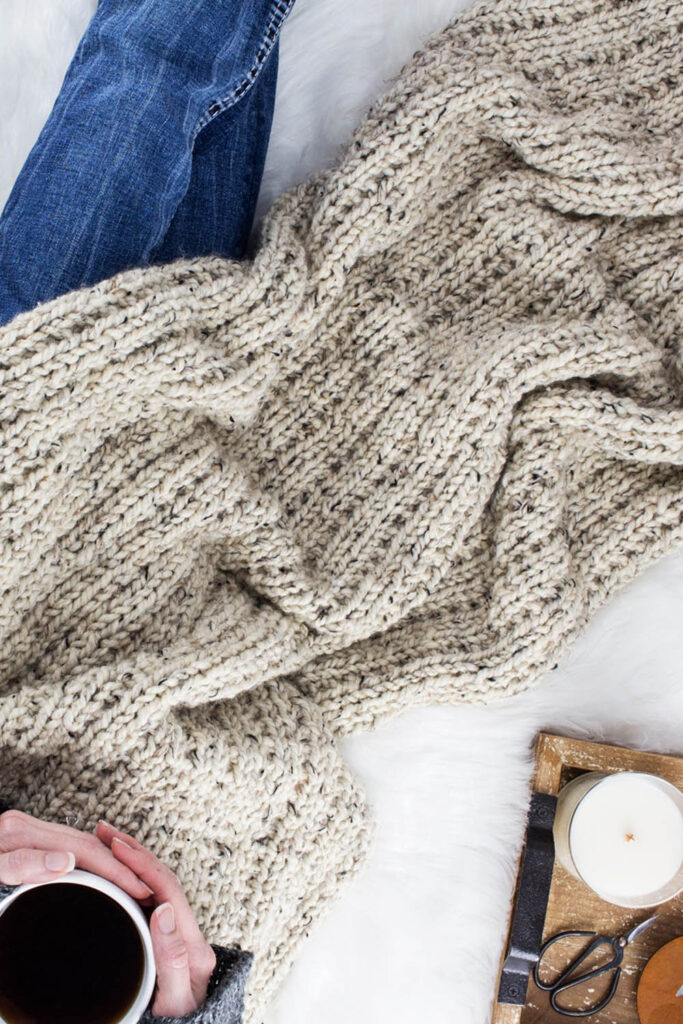 Model snuggled up in a cable knit blanket with a cup of coffee.