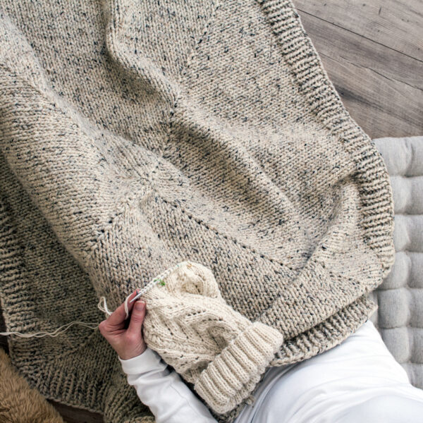 model snuggled up in a hand knit blanket knitting a hat