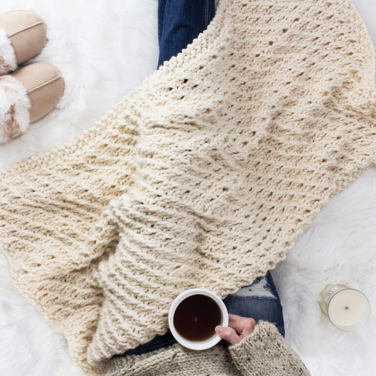 Model snuggled up in a cable knit blanket with a cup of coffee.