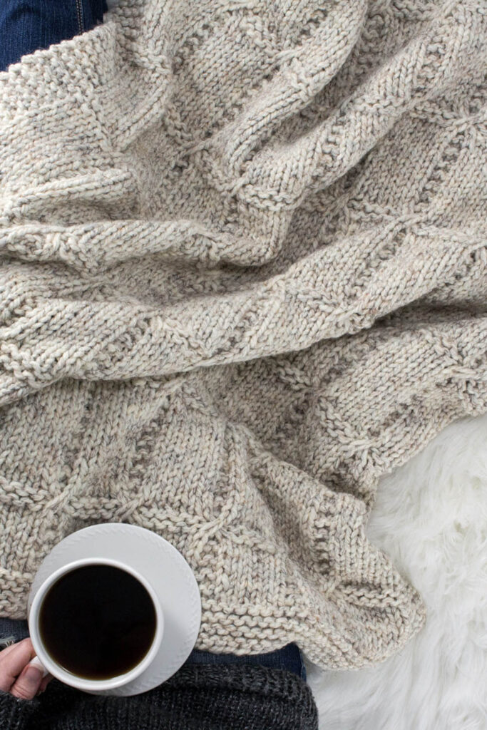 Model snuggled up in a beautiful hand knit blanket with a cup of coffee.