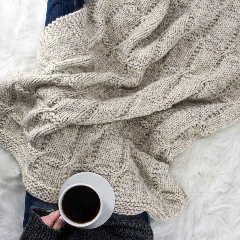 Model snuggled up in a beautiful hand knit blanket with a cup of coffee.