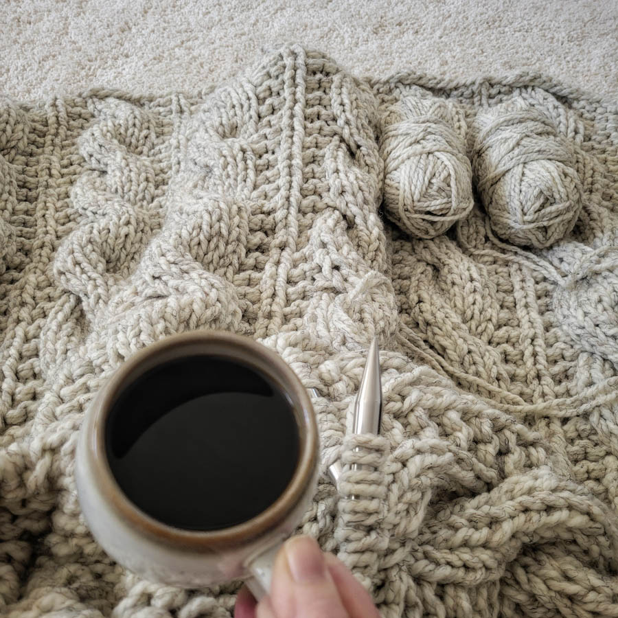 Model snuggled up in a cable knit blanket with a cup of coffee.