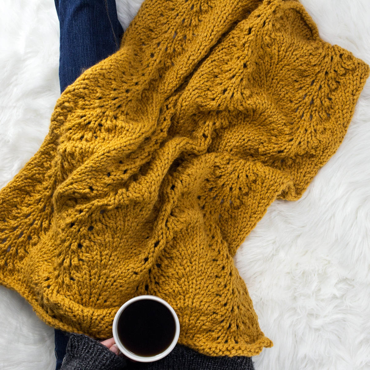 Model snuggled up in a cable knit blanket with a cup of coffee.