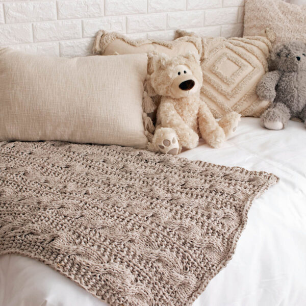 A cable knit blanket laying on a bed with a teddy bear.