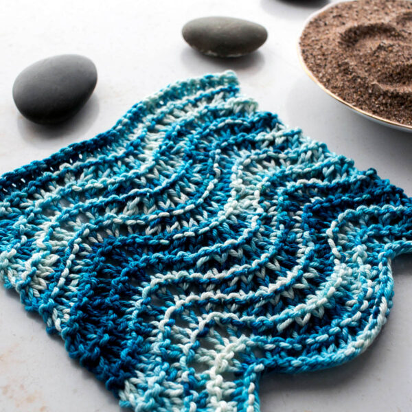 pic of a knitted lace dishcloth on a table with rocks & sand