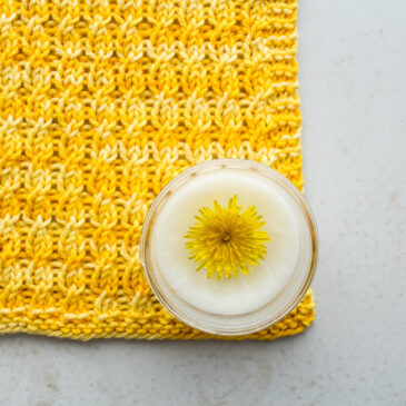 pic of a cable knitted dishcloth on a table with a jar of handmade dandelion lotion with a dandelion head in it.