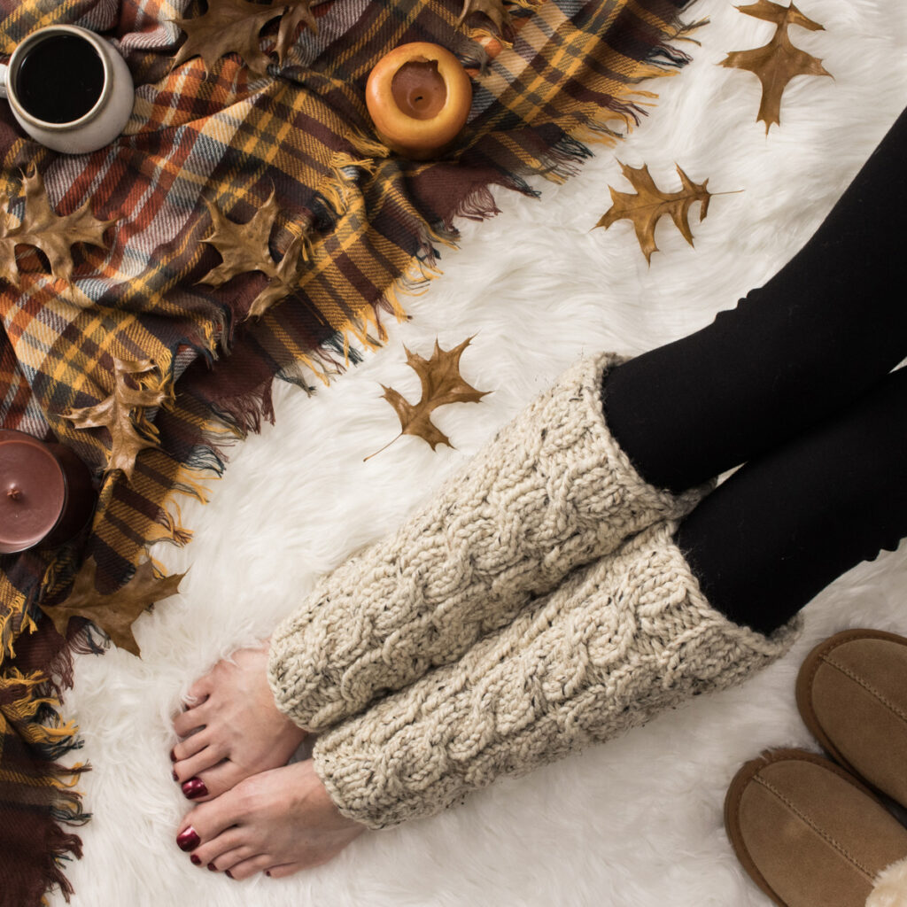 chunky cable knit leg warmers on a faux fur blanket in a cozy setting with plaid blanket, candles & coffee