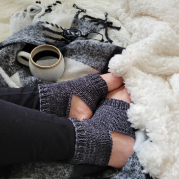 A cozy scene of knitted yoga socks on a faux fur blanket with a flannel blanket & cup of coffee.