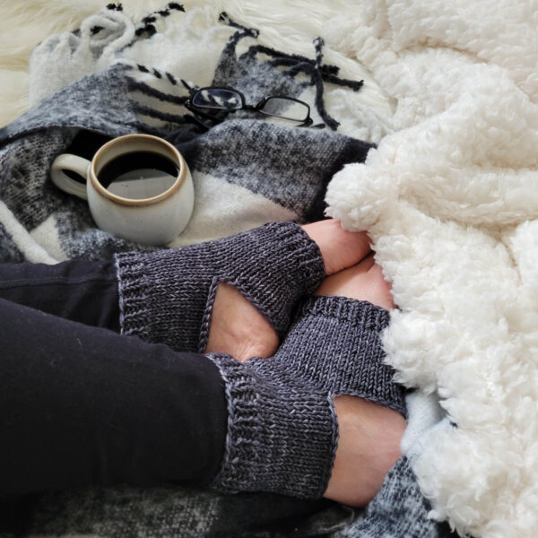 A cozy scene of knitted yoga socks on a faux fur blanket with a flannel blanket & cup of coffee.