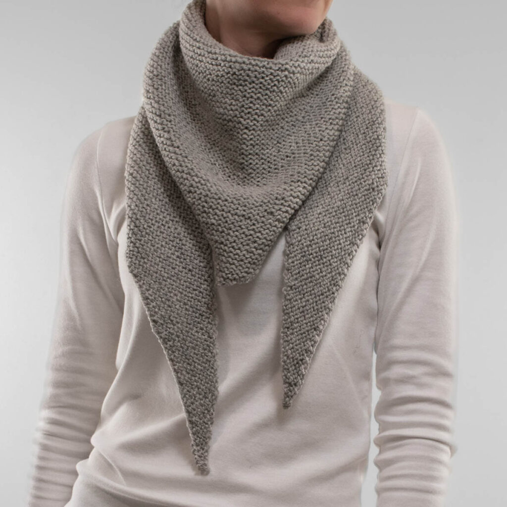 model wearing a handknit garter stitch skinny triangle scarf in soft grey, styled loosely around her neck over a white long-sleeve top.