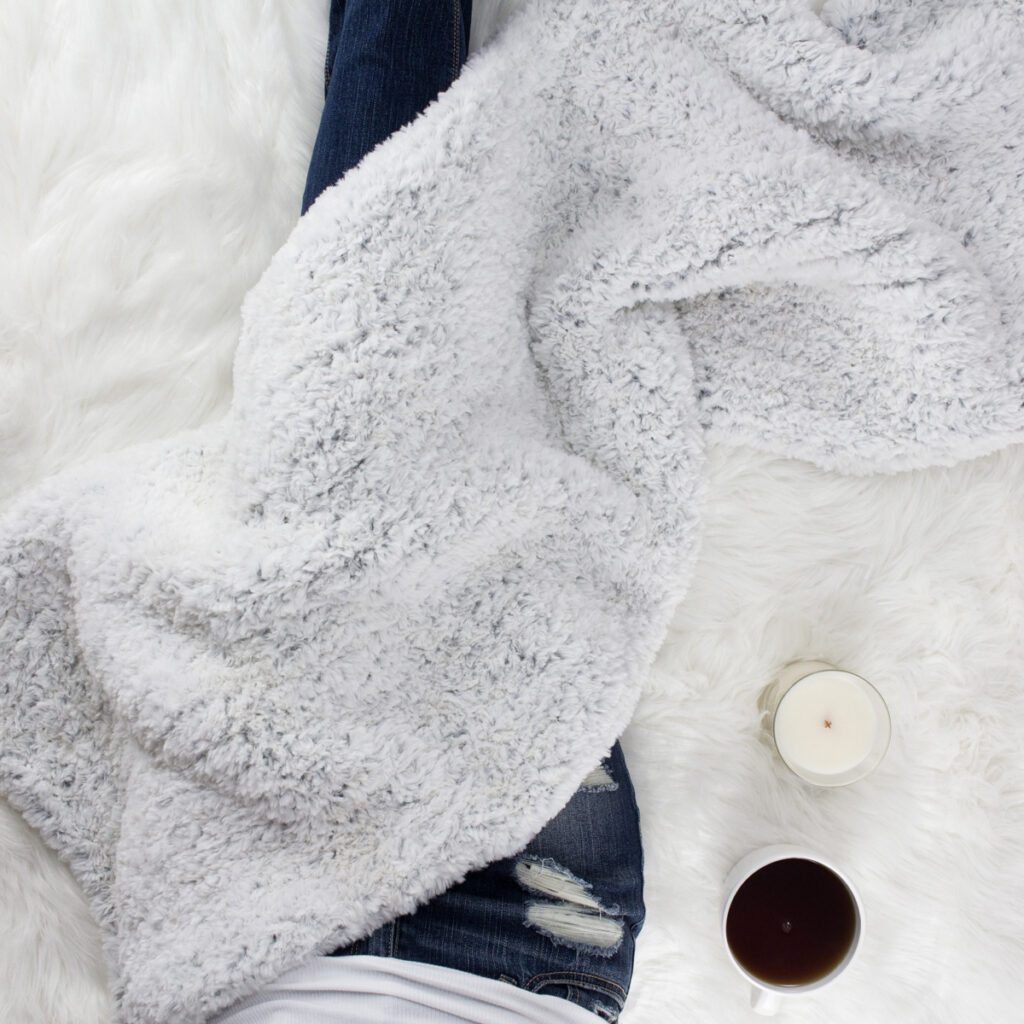 cozy scene of a rectangle shawl on a faux fur blanket with coffee & a candle.