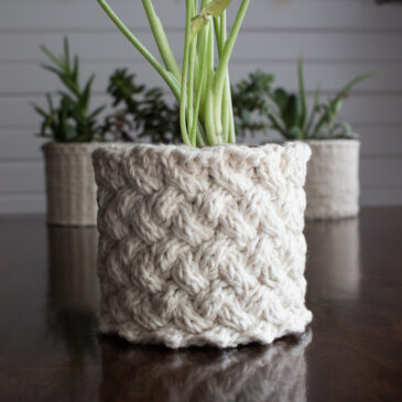 pic of a cable stitch knitted plant pot holder covering a pot with a plant in it