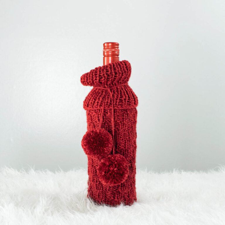 chevron knitted wine bottle cover with pom-poms displayed on a wine bottle.