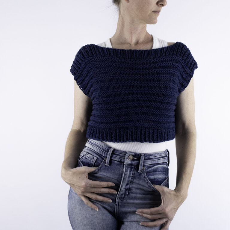 Woman wearing a hand-knit navy blue crop top poncho sweater made with DK weight yarn and ridge stitch, paired with light wash high-waisted jeans.
