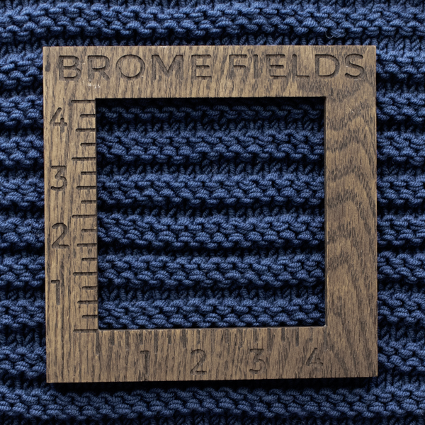 Knitting gauge swatch in navy blue ridge stitch with a wooden 4-inch gauge ruler marked "Brome Fields" placed on top to measure stitches and rows.