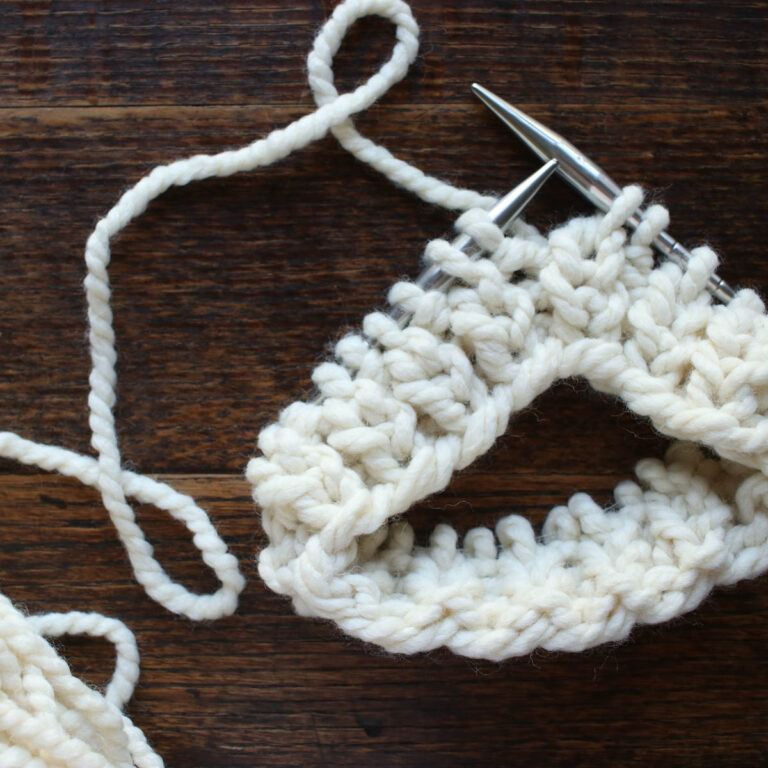 Close-up of Fisherman’s Rib Stitch being knit in the round on circular needles with chunky cream yarn.