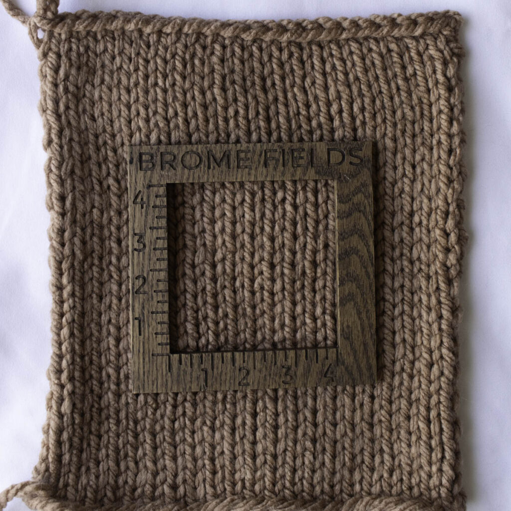 Square knit swatch in a neutral Paintbox Yarns Wool Mix Super Chunky, measured with a wooden 4-inch gauge tool, showing clean, even stockinette stitches with soft texture.