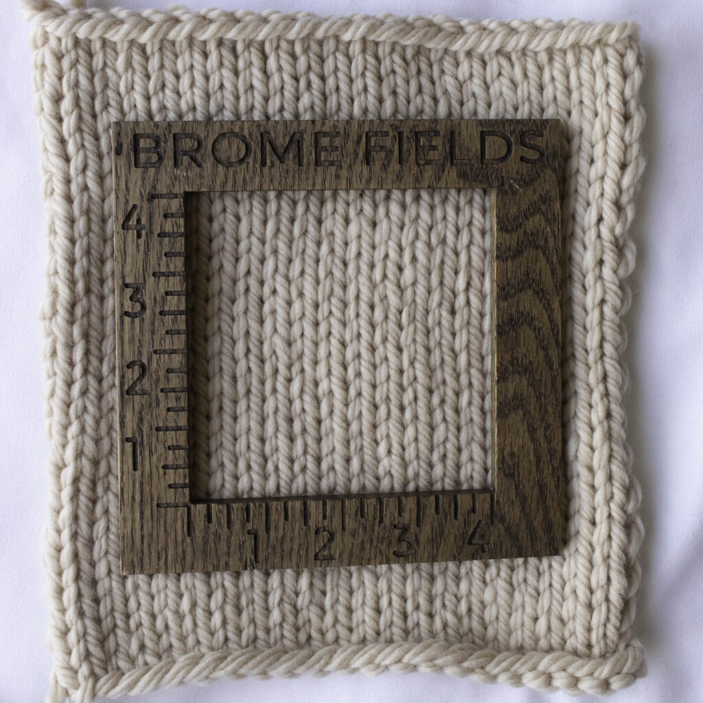 Close-up of a stockinette stitch knitting gauge swatch made with Rowan Big Wool in a soft cream color. The stitches are thick and fluffy with a matte finish, framed by a wooden Brome Fields gauge ruler.