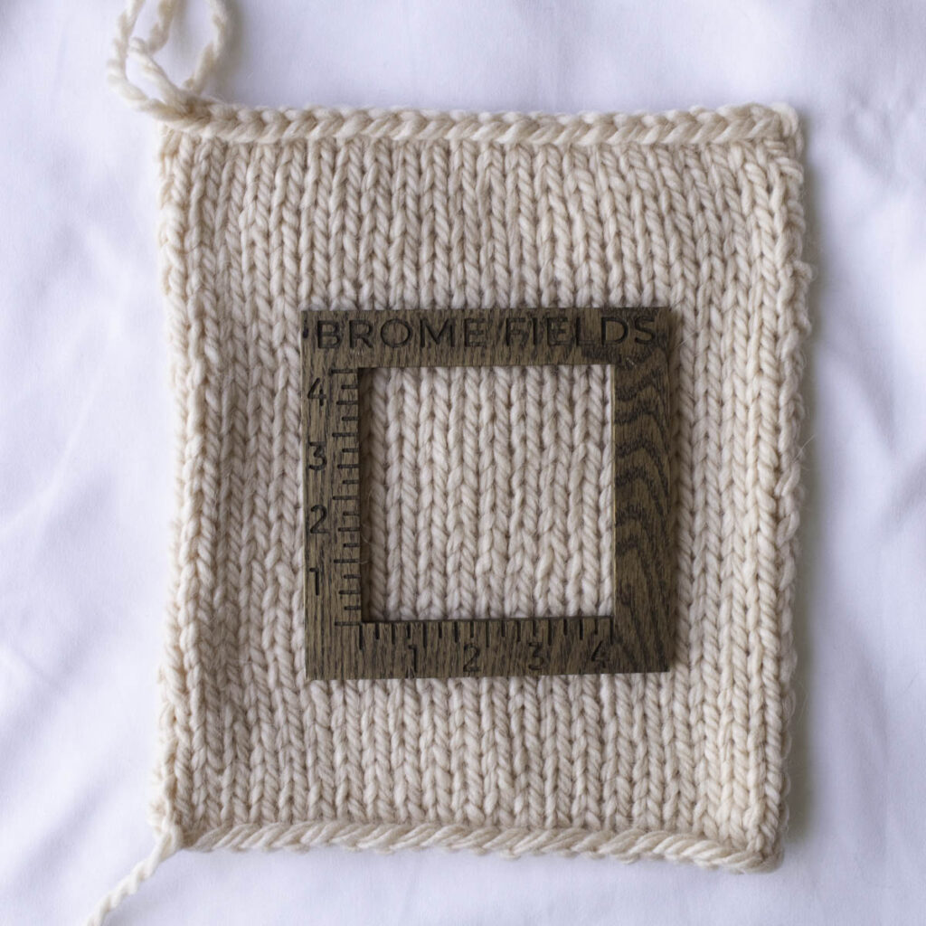 Swatch of Yarn Bee Effortless Alpaca Blend yarn in a soft, neutral cream color, shown in stockinette stitch with a wooden Brome Fields gauge tool on top.