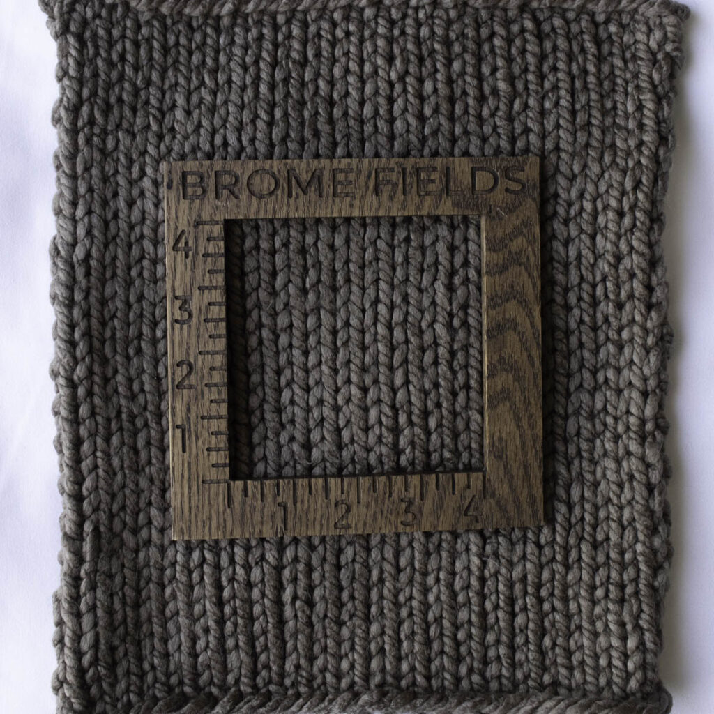 A close-up of a super bulky knitted swatch in Yarn Bee Dream Supreme Premium Micro-Acrylic yarn, shown with a Brome Fields wooden gauge ruler on top, highlighting stitch definition and soft texture in a warm taupe color.