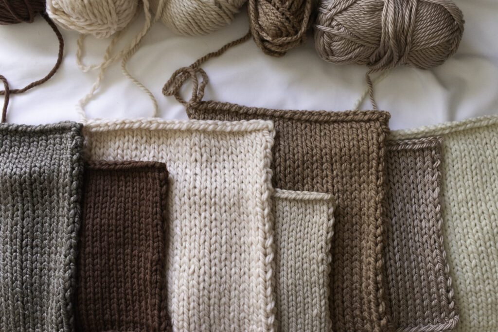 A lineup of seven hand-knit swatches in soft, earthy neutrals made with various super bulky yarns, including wool, alpaca, and acrylic blends. Each swatch is knit in stockinette stitch, showcasing different textures and tones. Yarn balls rest above the swatches on a white background.