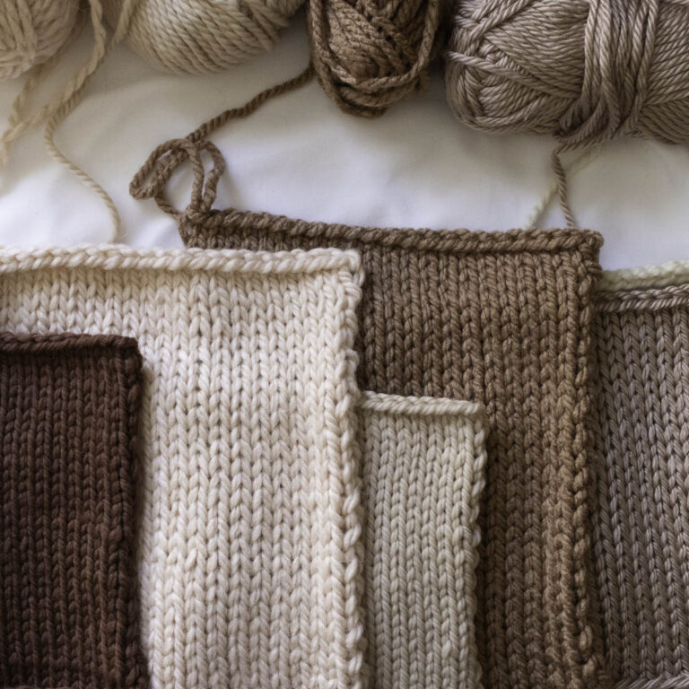 A lineup of seven hand-knit swatches in soft, earthy neutrals made with various super bulky yarns, including wool, alpaca, and acrylic blends. Each swatch is knit in stockinette stitch, showcasing different textures and tones. Yarn balls rest above the swatches on a white background.