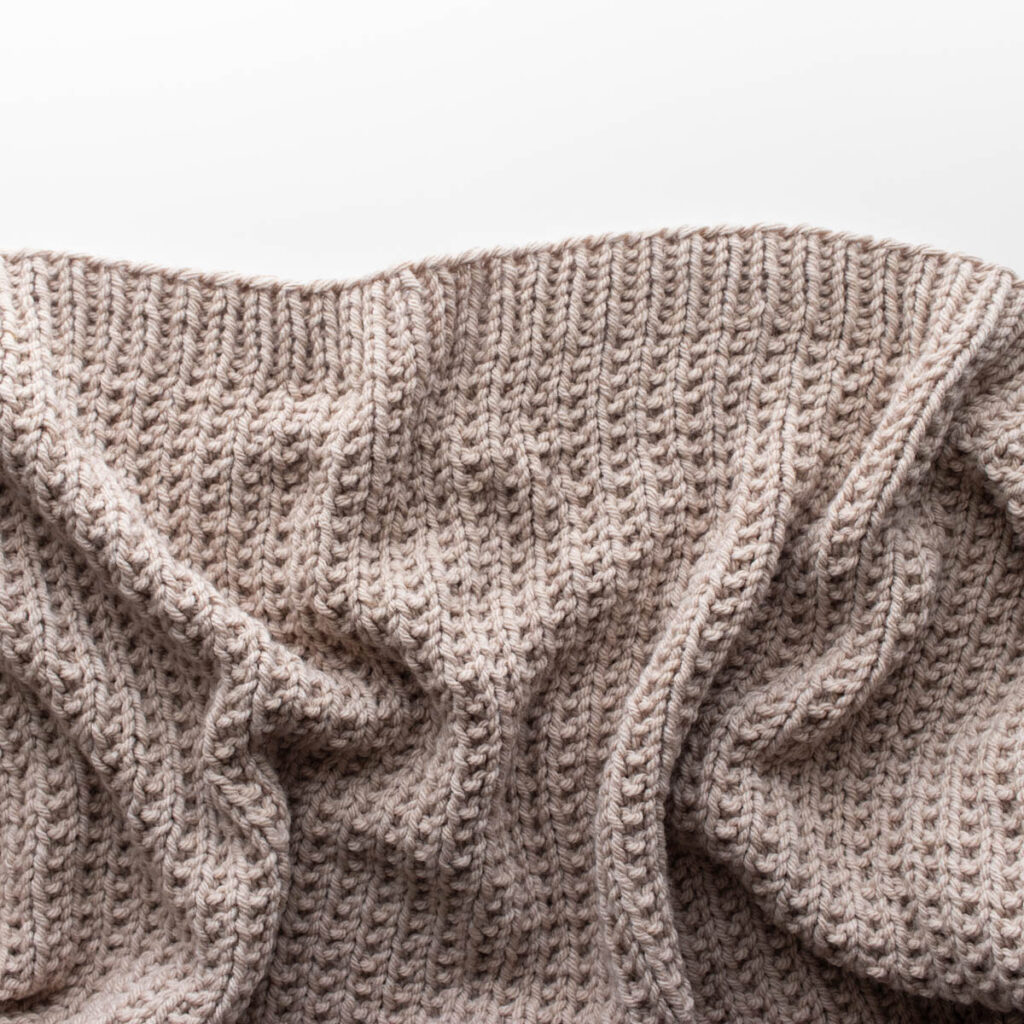 Close-up photo of a hand-knit beige throw blanket made with bulky yarn in a broken rib stitch pattern. The blanket is softly draped on a white surface, showing its cozy texture and reversible design with beautiful stitch definition.