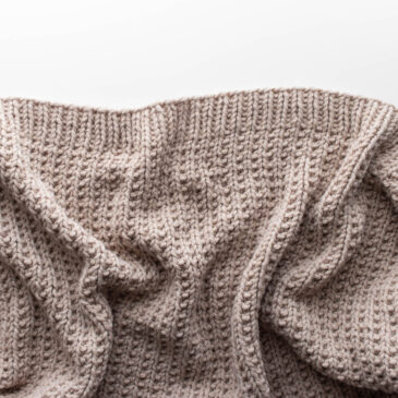 Close-up photo of a hand-knit beige throw blanket made with bulky yarn in a broken rib stitch pattern. The blanket is softly draped on a white surface, showing its cozy texture and reversible design with beautiful stitch definition.