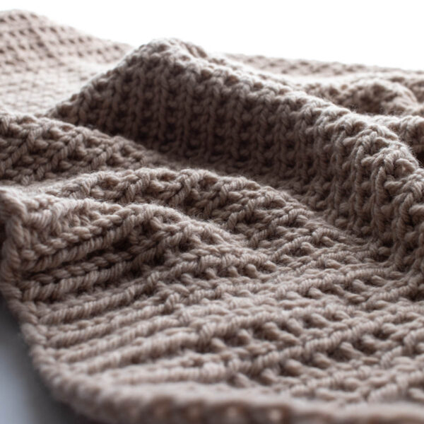 Close-up photo of a hand-knit beige throw blanket made with bulky yarn in a broken rib stitch pattern. The blanket is softly draped on a white surface, showing its cozy texture and reversible design with beautiful stitch definition.