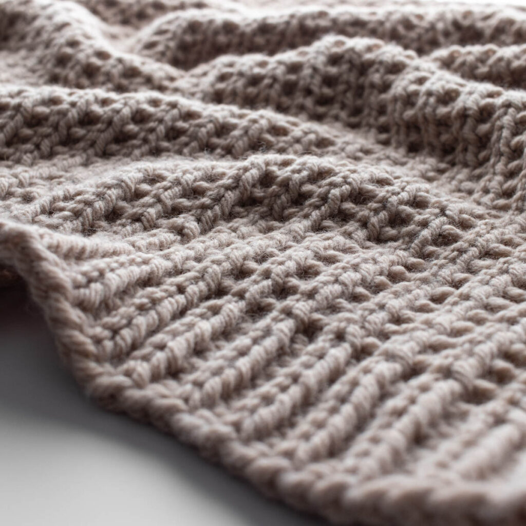 Close-up photo of a hand-knit beige throw blanket made with bulky yarn in a broken rib stitch pattern. The blanket is softly draped on a white surface, showing its cozy texture and reversible design with beautiful stitch definition.