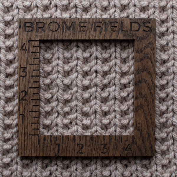 Close-up photo of a bulky yarn swatch in a broken rib stitch pattern, measured with a wooden 4-inch gauge ruler engraved with “Brome Fields.” The yarn is soft beige, showing clear stitch definition and beautiful texture.