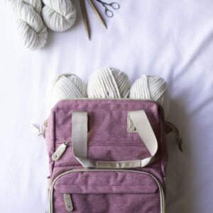 Mauve travel knitting bag filled with chunky white yarn on a white background, with wooden knitting needles and small scissors placed above it.