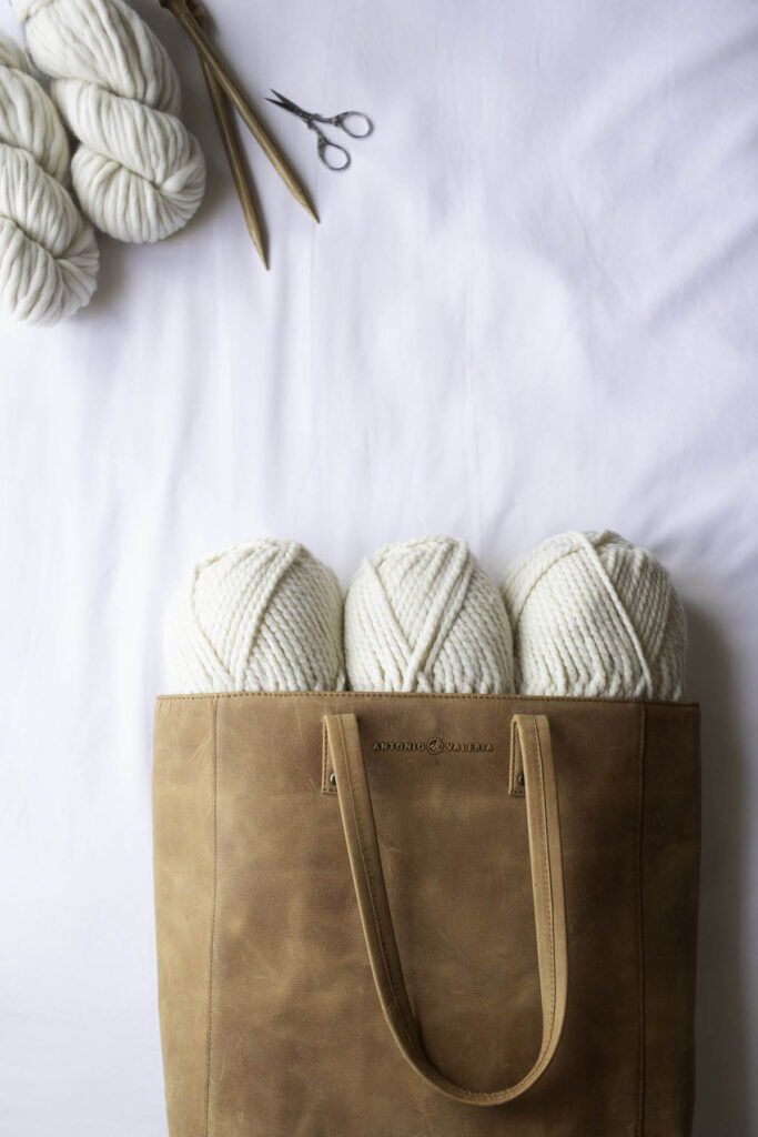 Rustic cognac leather knitting bag filled with soft white yarn and knitting needles, styled with yarn skeins and scissors on a white backdrop.