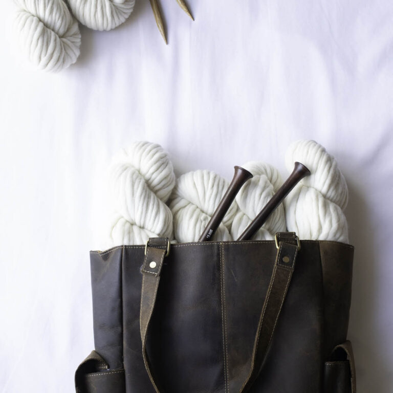 Knitting bag made of distressed leather, filled with bulky cream yarn, styled for a knitting-themed flat lay.