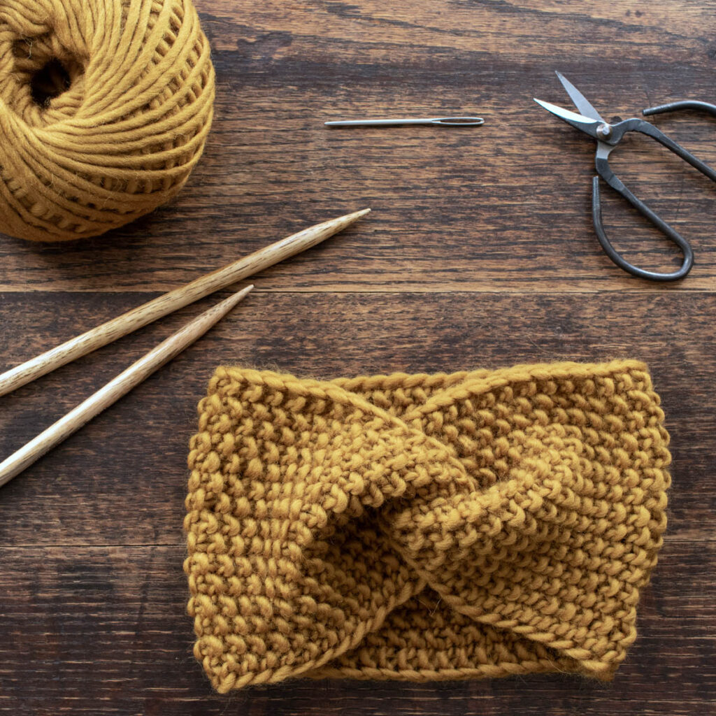 Hands holding a chunky seed stitch twist knit headband on a wooden table with yarn, knitting needles, and scissors