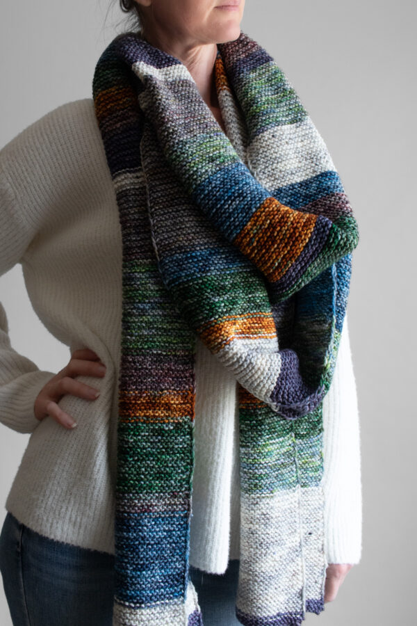Woman wearing an extra long colorful striped garter stitch scarf in hand-dyed multicolor yarn, draped around her neck.