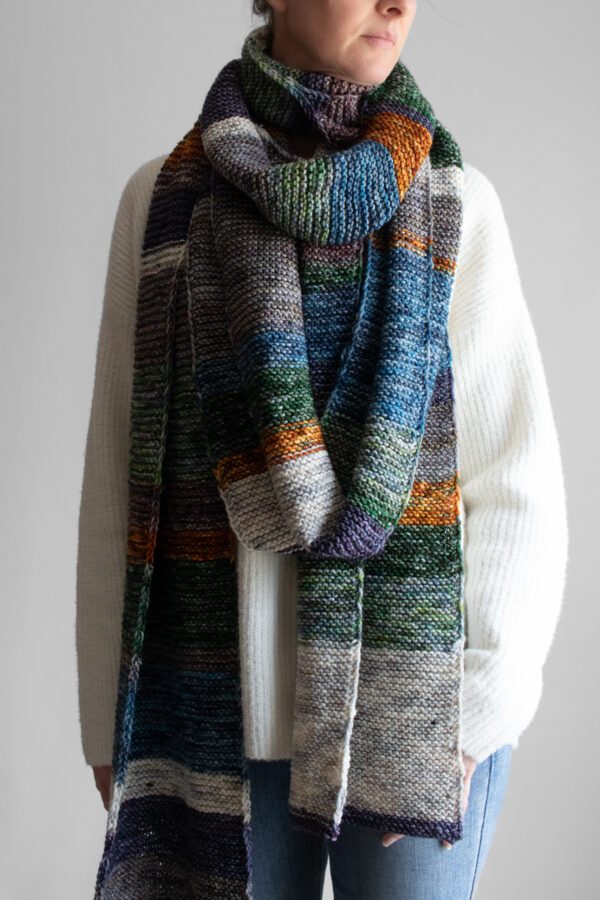 Woman wearing an extra long colorful striped garter stitch scarf in hand-dyed multicolor yarn, draped around her neck.