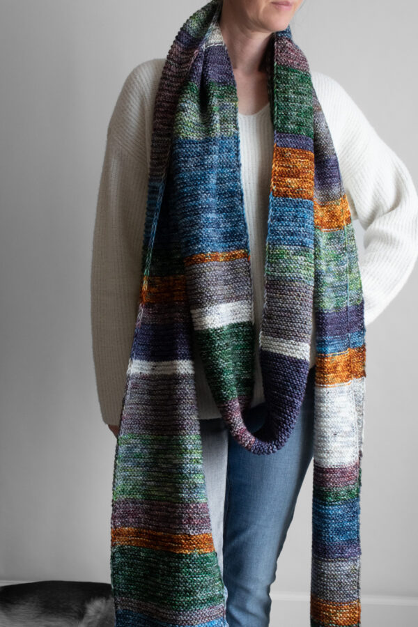 Woman wearing an extra long colorful striped garter stitch scarf in hand-dyed multicolor yarn, draped around her neck.